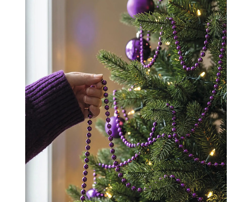 Eine Hand dekoriert einen Christbaum mit einer lila Perlengirlande, Christbaumkugeln und warmweißer Beleuchtung.