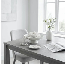 Helles Esszimmer mit grauem Holztisch, hellgrauem Polsterstuhl, weißer Dekoschale mit Perlenketten, weißer Vase mit Blumen und offenem Magazin.