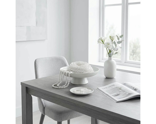 Helles Esszimmer mit grauem Holztisch, hellgrauem Polsterstuhl, weißer Dekoschale mit Perlenketten, weißer Vase mit Blumen und offenem Magazin.