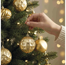 Eine Hand hängt eine goldene Weihnachtskugel mit geometrischem Muster an einen grünen Weihnachtsbaum mit warmen Lichtern.