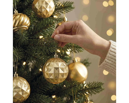 Eine Hand hängt eine goldene Weihnachtskugel mit geometrischem Muster an einen grünen Weihnachtsbaum mit warmen Lichtern.
