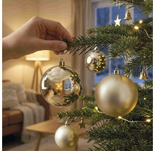 Eine Hand hängt eine glänzende Weihnachtskugel an einen geschmückten Weihnachtsbaum mit Lichtern in einem hellen Wohnzimmer.