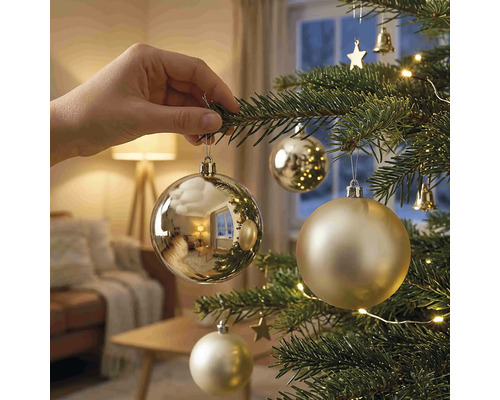 Eine Hand hängt eine glänzende Weihnachtskugel an einen geschmückten Weihnachtsbaum mit Lichtern in einem hellen Wohnzimmer.