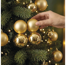 Hand dekoriert einen Christbaum mit matten und glänzenden Christbaumkugeln und einer Lichterkette.