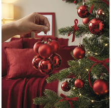 Eine Hand hält ein Set roter Weihnachtskugeln vor einem festlich geschmückten Weihnachtsbaum mit Schleifen und Lichterkette.