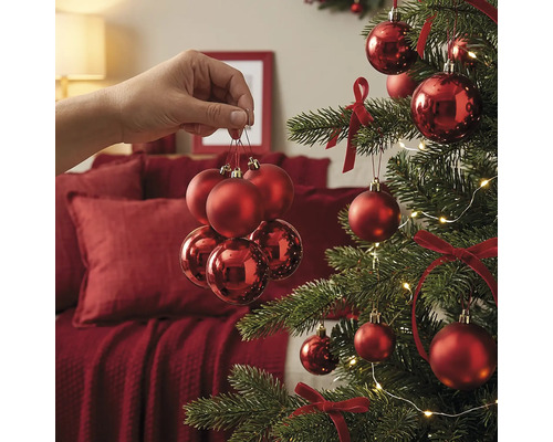 Eine Hand hält ein Set roter Weihnachtskugeln vor einem festlich geschmückten Weihnachtsbaum mit Schleifen und Lichterkette.