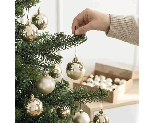 Eine Hand hängt eine champagnerfarbene Weihnachtskugel an einen grünen Weihnachtsbaum. Im Hintergrund steht eine offene Schachtel mit Kugeln.