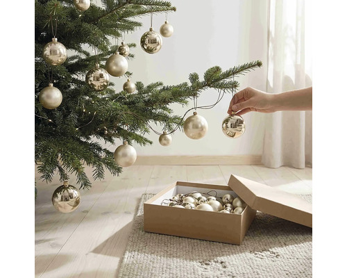 Eine Hand hängt eine champagnerfarbene Weihnachtskugel an einen Tannenbaum. Am Boden steht ein Karton mit Christbaumkugeln.