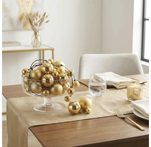 Festlich gedeckter Holztisch mit goldenem Tischläufer, einer Glasschale voller goldener Weihnachtskugeln, einer brennenden Kerze und Besteck.
