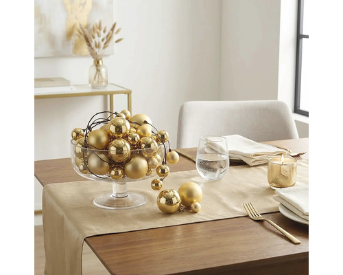 Festlich gedeckter Holztisch mit goldenem Tischläufer, einer Glasschale voller goldener Weihnachtskugeln, einer brennenden Kerze und Besteck.