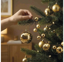 Hand hängt eine goldene Weihnachtskugel an einen Tannenbaum, der mit goldenem Christbaumschmuck und einer Lichterkette dekoriert ist.
