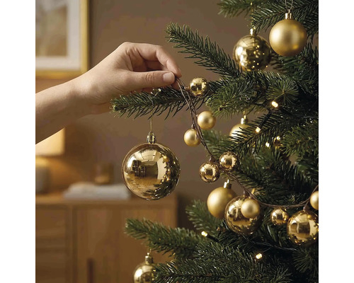 Hand hängt eine goldene Weihnachtskugel an einen Tannenbaum, der mit goldenem Christbaumschmuck und einer Lichterkette dekoriert ist.