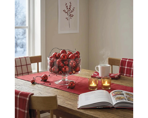 Festlich gedeckter Holztisch mit rotem Läufer, einer Glasschale voller Weihnachtskugeln, Teelichtern und einem Kochbuch.