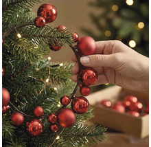 Hand beim Dekorieren eines Christbaums mit einer Girlande aus roten Weihnachtskugeln verschiedener Größen und warmweißen Lichtern.