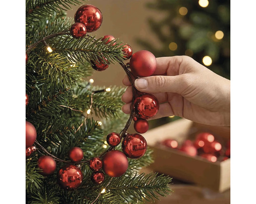 Hand beim Dekorieren eines Christbaums mit einer Girlande aus roten Weihnachtskugeln verschiedener Größen und warmweißen Lichtern.