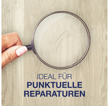 Hand hält eine Lupe über eine Holzoberfläche mit einem Kratzer, um punktuelle Reparaturen zu veranschaulichen.