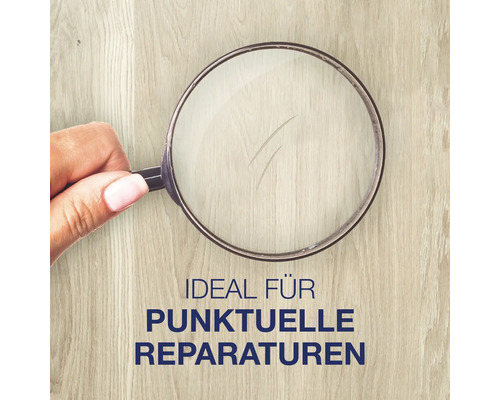 Hand hält eine Lupe über eine Holzoberfläche mit einem Kratzer, um punktuelle Reparaturen zu veranschaulichen.