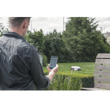 Mann mit Smartphone im Garten, im Hintergrund ein Rasenmähroboter
