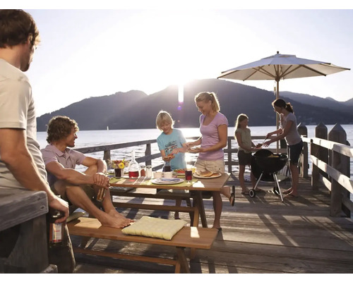 Familie grillt auf einem Holzsteg am See mit Grill, Sonnenschirm und Picknicktisch