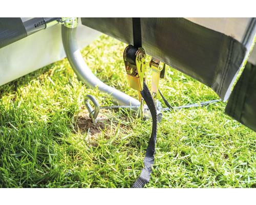 Detailaufnahme einer Plane, die mit einem Ratschengurt und einem Haken im Boden befestigt ist. Der Boden ist mit Gras bedeckt.
