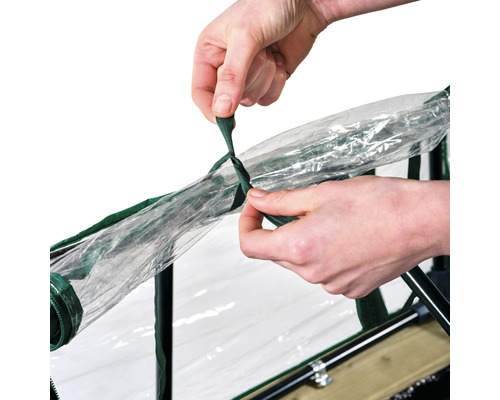 Eine Person befestigt eine transparente Folie mit einem grünen Band an einem Frühbeet.