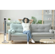 Ein Turmventilator steht in einem hellen Wohnzimmer neben einem Sofa, auf dem eine entspannte Frau sitzt.