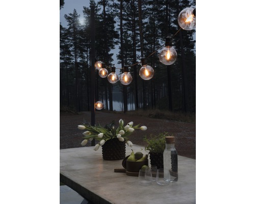 10er LED Lichterkette Retro 4,50 m Lichtfarbe bernstein dimmbar Beleuchteter Außenbereich mit Lichterkette, Tisch, Blumen und Getränken vor Waldkulisse