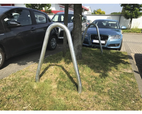 Zwei Fahrradständer aus Metall auf einer Grünfläche zwischen parkenden Autos.