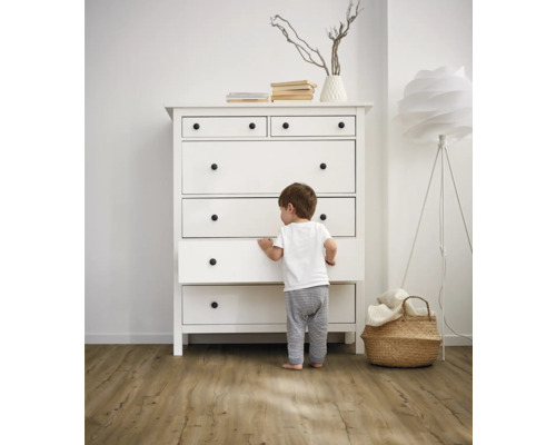 Ein Kind steht vor einer weißen Kommode mit Schubladen und runden Griffen in einem hellen Raum.