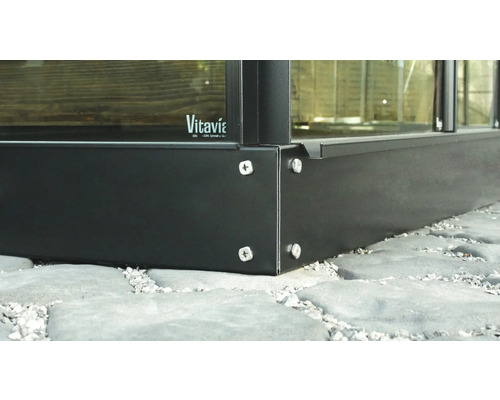 Detailansicht eines Gewächshausfundaments aus Metall mit Schraubenbefestigung auf Steinfliesenboden. Vitavia Logo.