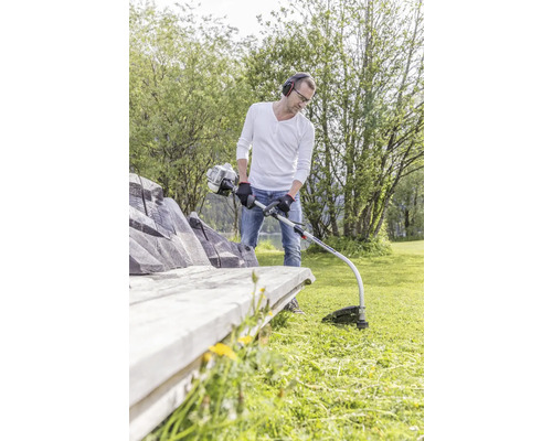 Mann schneidet Gras mit einem Rasentrimmer neben einer Bank in einem Garten.