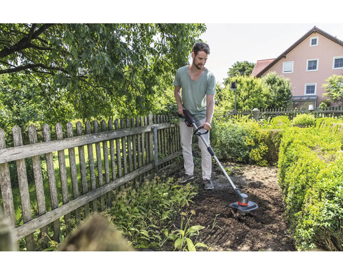 Ein Mann benutzt einen Rasentrimmer in einem Garten mit Zaun und Büschen.