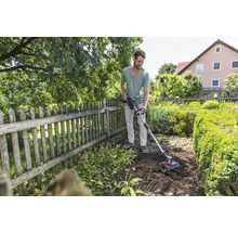 Ein Mann benutzt einen Rasentrimmer in einem Garten mit Holzzaun und Büschen.