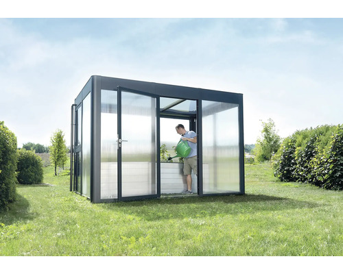 Mann gießt Pflanzen in einem Gewächshaus mit geöffneten Türen auf einer Wiese.