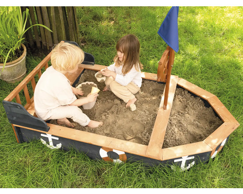 Zwei Kinder spielen in einem Sandkasten in Bootsform mit Steuerrad und Segel im Garten.