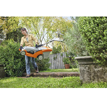 Eine Frau trägt einen Rasenmäher durch einen Garten.