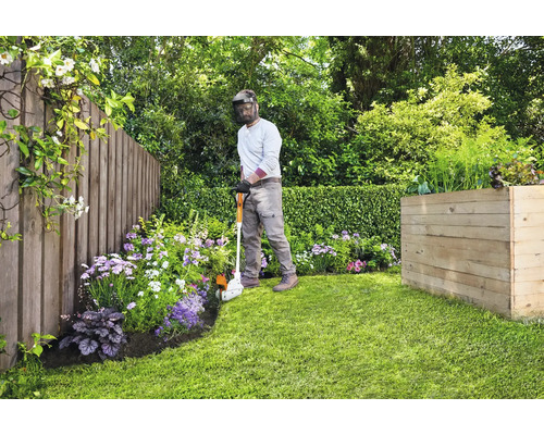 Gärtner schneidet Rasenkanten mit einem Kantenschneider in einem Garten mit Holzzaun und Hochbeet