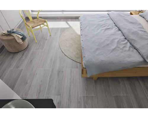 Schlafzimmer mit Bett, Stuhl, Hocker, Teppich und grauen Bodenfliesen in Holzoptik