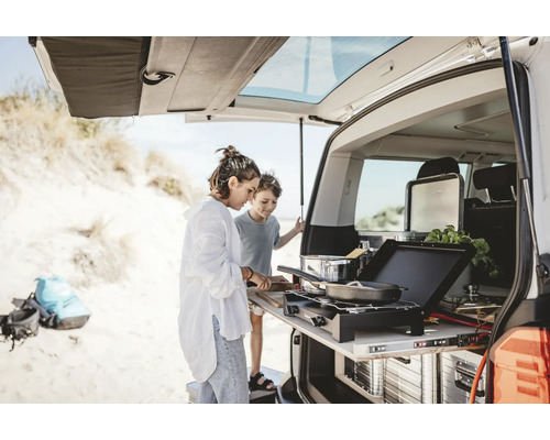Zwei Personen kochen auf einem Auszug-Kochfeld am Heck eines Fahrzeugs am Strand.