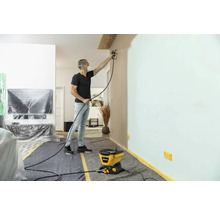 Mann streicht Wand mit Farbsprühsystem in einem Wohnraum, der mit Abdeckmaterialien geschützt ist.