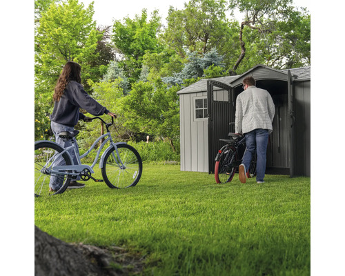 Gartenszene mit Gerätehaus und zwei Personen mit Fahrrädern auf Rasenfläche