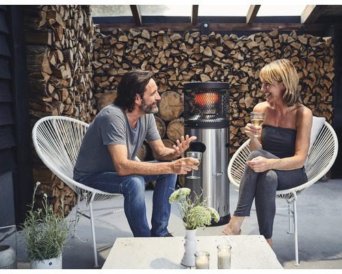 Szene mit einem Paar, das auf Sesseln neben einem Heizpilz sitzt und sich unterhält, im Hintergrund gestapeltes Holz.
