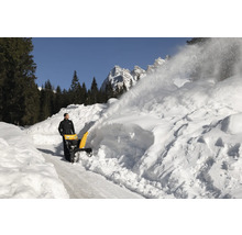 Eine Schneefräse räumt eine verschneite Straße frei, im Hintergrund sind Bäume und Berge zu sehen.
