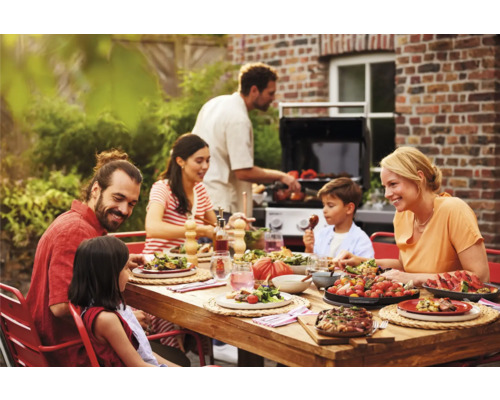 Familie genießt Mahlzeit am Holztisch im Garten mit Grill im Hintergrund