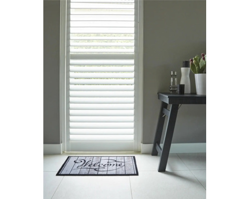 Szene mit Türjalousien, Fußmatte mit der Aufschrift Willkommen und Beistelltisch mit Dekorationsartikeln