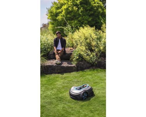 Szene mit einem Rasenmähroboter auf einer Rasenfläche und einer Person, die auf Felsen sitzt