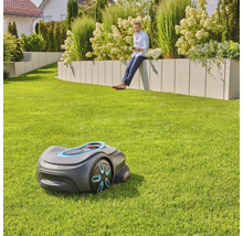 Szene mit einem Rasenmähroboter auf einer Rasenfläche und einem Mann, der auf einer Gartenmauer sitzt und ein Mobiltelefon benutzt