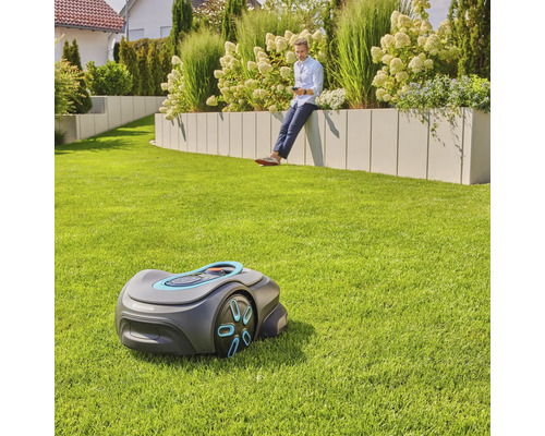 Szene mit einem Rasenmähroboter auf einer Rasenfläche und einem Mann, der auf einer Gartenmauer sitzt und ein Mobiltelefon benutzt
