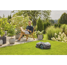 Mann entspannt im Garten mit Rasenmähroboter, Pflanzen und Gartenmöbeln