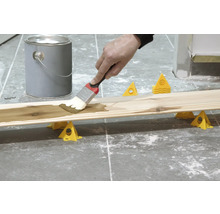 Holzbrett wird mit einem Pinsel gestrichen, Farbdose und Malereihilfen sind sichtbar.